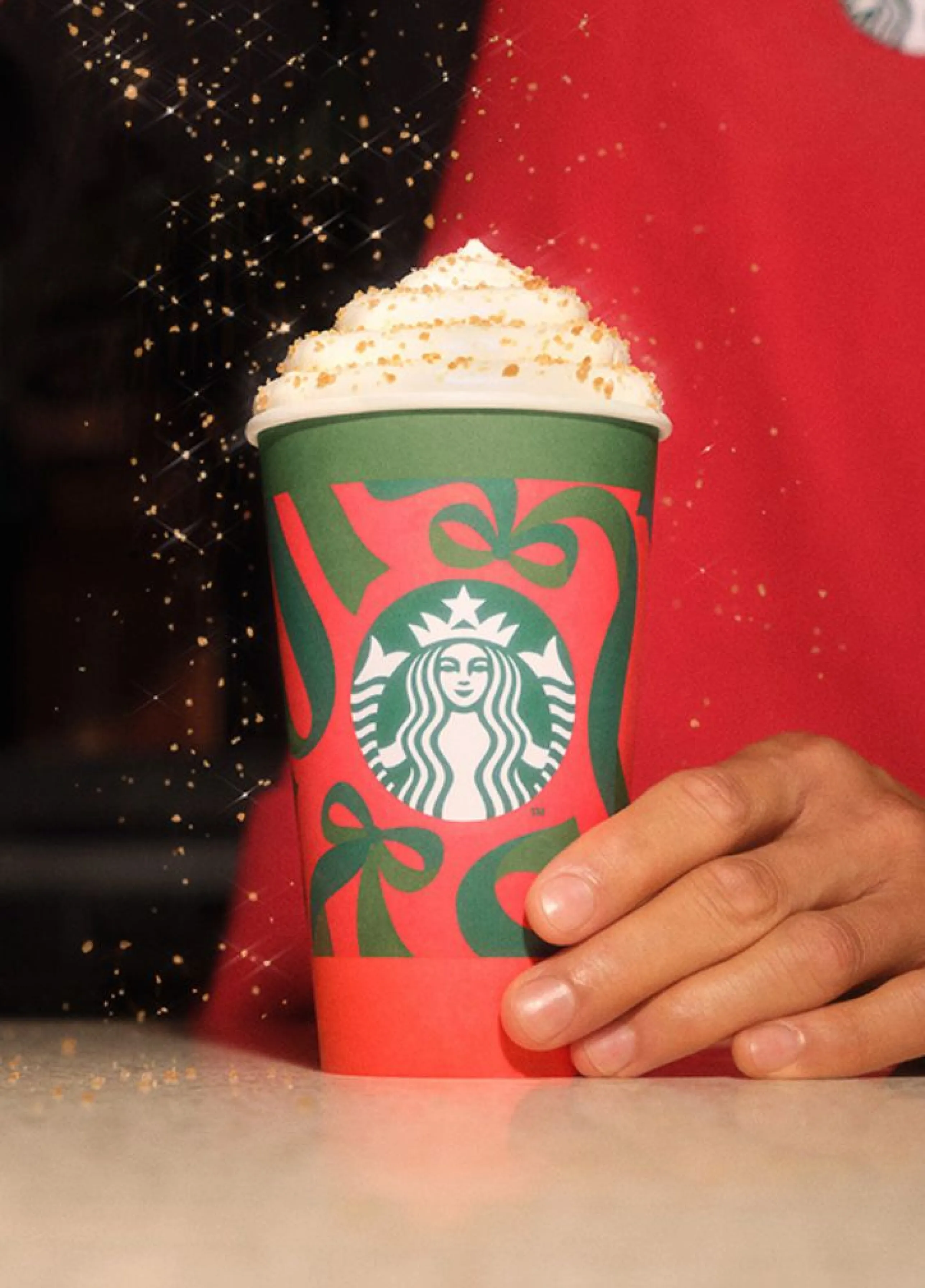 Kelímek Starbucks s vánočním motivem s nápojem se šlehačkou, na kterou sype barista cukrovou posypku