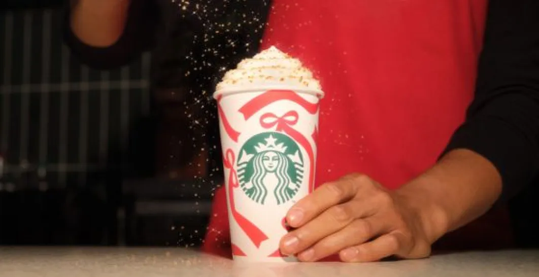A Starbucks cup with a Christmas motif containing a drink topped with whipped cream, onto which the barista sprinkles sugar sprinkles