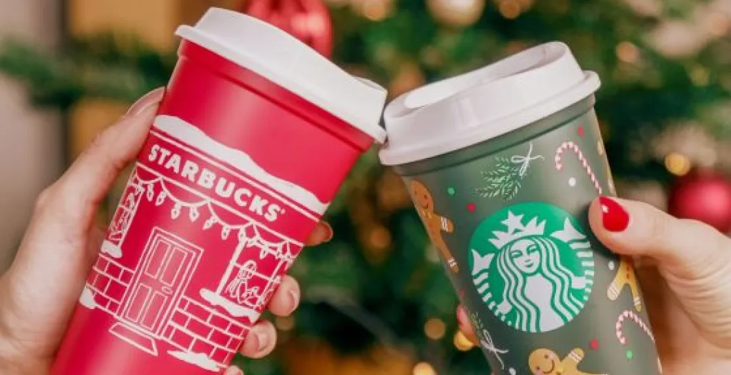 One hand holds a reusable cup with a red and white Christmas motif and a white lid, and the other hand holds a reusable green mug with gingerbread cookies and a white lid